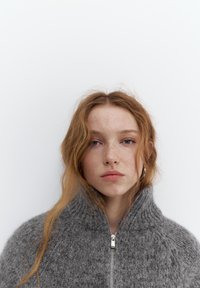 Młoda kobieta o rudych włosach i piegach, ubrana w szary sweter z wysokim kołnierzem i zamkiem, na jednolitym białym tle.