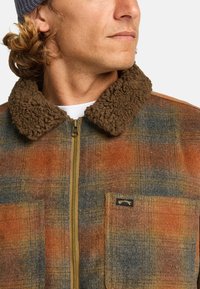 Bruine en oranje geruite jas met een zachte bruine kraag, ritssluiting en twee voorzakken met een merklabel.