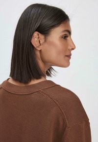 Brauner Strickpullover mit rundem Halsausschnitt und Raglanärmeln, aus strukturiertem Stoff und mit lockerer Passform, von der Seite gezeigt.