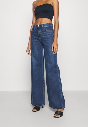 Donna che indossa jeans a vita alta a gamba larga blu scuro, top nero senza spalline e sandali neri con tacco su pavimento grigio e sfondo bianco.