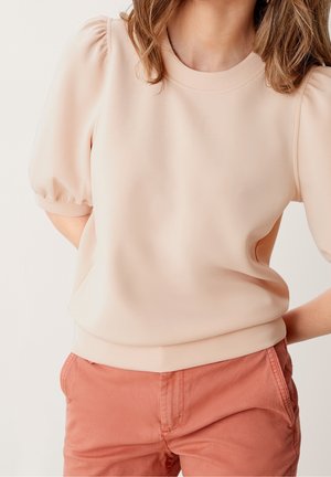 Kobieta ubrana w jasnobeżową bluzkę z krótkimi bufkami i koralowo-różowe spodnie, z rękami ułożonymi za plecami.