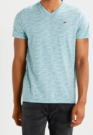 Man wearing a light blue heathered V-neck t-shirt with a small red bird logo on the left chest and dark pants, standing against a white background.