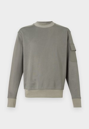 Grå genser med lange ermer, rund hals og en lomme på venstre erme. Myk tekstur, ribbestrikkede mansjetter og kantdetaljer.