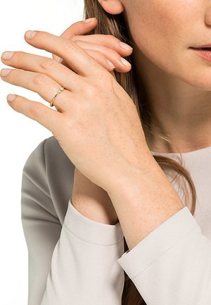 Goldring mit einem kleinen zentralen Stein, getragen an einer Hand mit heller Haut und natürlich aussehenden Nägeln, vor einem sanften grauen Hemd.