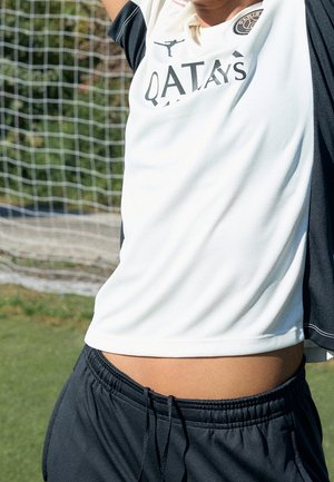 Personne portant un maillot de sport blanc et un short noir, levant les bras devant un but de football sur un terrain herbeux.
