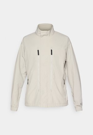 Giacca di un beige chiaro con colletto alto, tasche frontali con zip, polsini elasticizzati e una texture liscia e leggera.