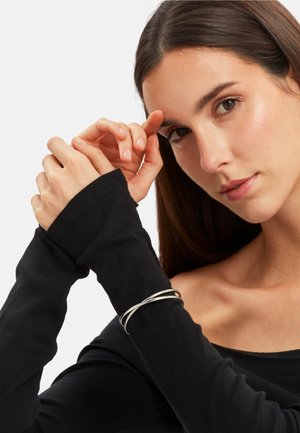 Sort langærmet bluse med tætsiddende manchetter, der viser et sølv multi-strengs armbånd på håndleddet. Glat stof med en let glans.