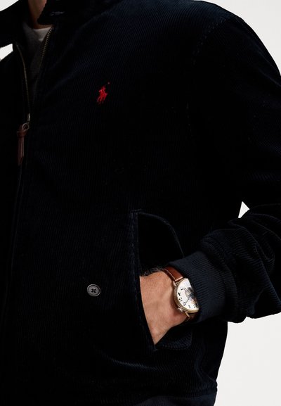 Giacca di velluto a coste blu marina con zip frontale, polsini a coste e tasche. Presenta un piccolo logo rosso e un orologio in pelle marrone al polso.
