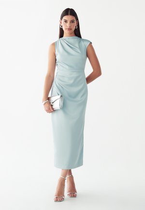 Donna che indossa un vestito di raso azzurro chiaro senza maniche, tiene una pochette argentata e indossa sandali argentati con tacco alto e cinturini su uno sfondo semplice.