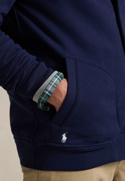 Άτομο που φοράει σκούρο μπλε φούτερ με κουκούλα, με το χέρι στην τσέπη, φορεμένο πάνω από πράσινο καρό πουκάμισο και γκρι μανίκι, με μπεζ παντελόνι ορατό στη μέση.