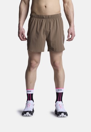 Man draagt bruine sportshorts, zwarte en roze sokken en witte hardloopschoenen, staand tegen een effen witte achtergrond.