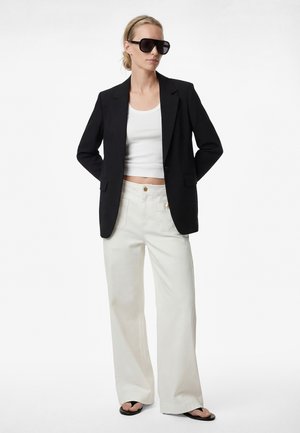 Mujer con blazer negro, top corto blanco, pantalones anchos blancos, chanclas negras, gafas de sol negras y una diadema, de pie con las manos detrás de la espalda.