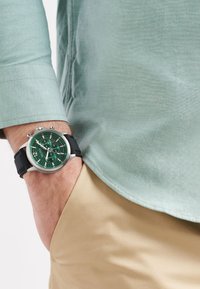 Montre argentée avec un cadran texturé vert, bracelet en silicone noir, portée sur un poignet à côté d'une chemise vert menthe et d'un pantalon beige.