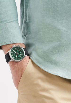 Orologio argento con quadrante verde testurizzato, cinturino in silicone nero, indossato al polso insieme a una camicia verde menta e pantaloni beige.