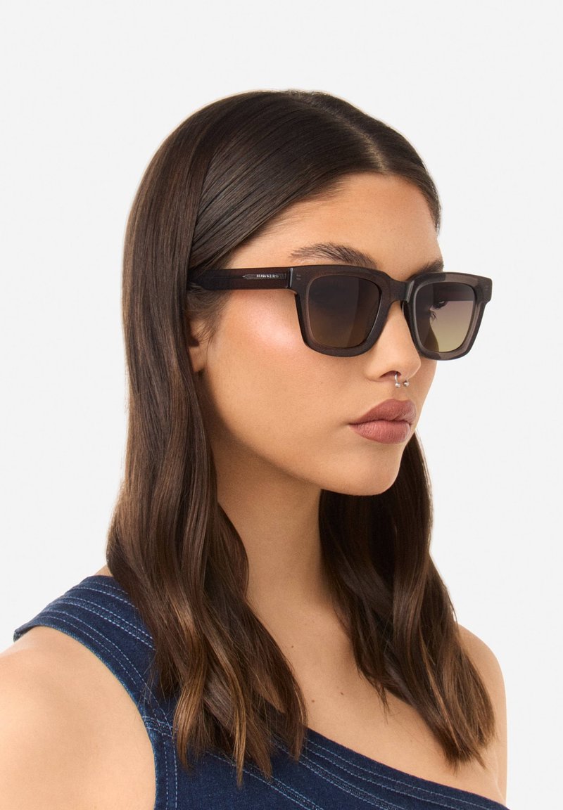 Occhiali da sole con una robusta montatura nera opaca e lenti marroni sfumate, caratterizzati da forme angolari e un branding sottile sulla gamba.