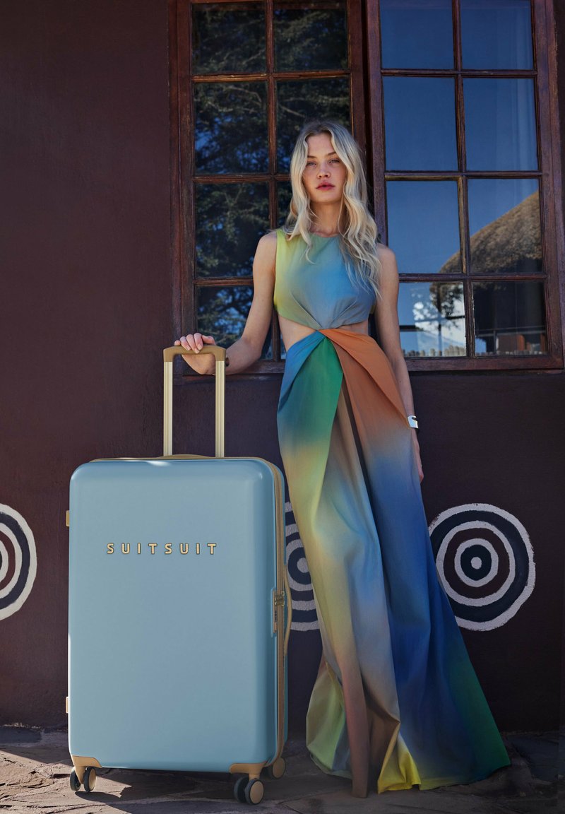 Harde koffer in pastelblauw met gouden hardware, vier wielen en een uitschuifbare handgreep. Opvallend door de gladde afwerking en het merkembleem.