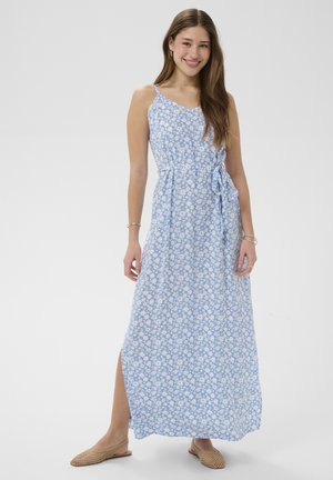Ung kvinde iført en ærmeløs lyseblå maxikjole med hvidt blomsterprint, sidelommer og beige vævede slip-on sko, smilende til kameraet.