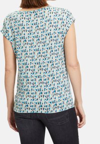 Tričkot s krátkým rukávem s světlou modrou barvou pozadí a geometrickým vzorem v tmavších odstínech modré a hnědé, vyrobené z měkké a lehké tkaniny.