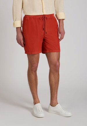 Mann trägt rote Shorts mit Kordelzug, weiße Turnschuhe und ein beiges Langarmhemd mit hochgekrempelten Ärmeln, steht vor einem schlichten Hintergrund.