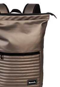 Rugzak met een gladde, beige buitenkant, voorzien van een gevoerde voorzak, zwarte ritsen en een prominente logolabel aan de onderkant.