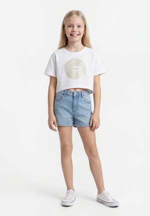 Κορίτσι που χαμογελά με μακριά ξανθά μαλλιά, ντυμένο με λευκό crop T-shirt με αποτύπωμα «Moschino» σε κύκλο και ανοιχτό μπλε τζιν σορτς.