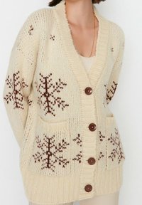 Beige gestrickte Strickjacke mit Schneeflockenmustern in Braun, mit V-Ausschnitt, aufgesetzten Taschen und vier braunen Knöpfen an der Vorderseite.