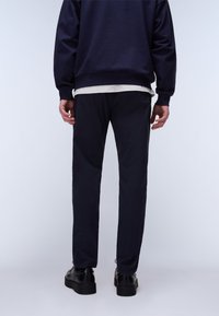 Mand iført sorte bukser, sorte sko, navyblå sweatshirt og grå undertrøje, stående med ryggen imod mod en ensfarvet hvid baggrund.