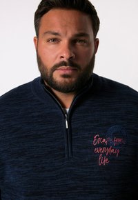 Marinblå zip-up tröja med strukturerad stickning, med röd broderad text och en cirkulär logotypdetalj på vänster bröst.