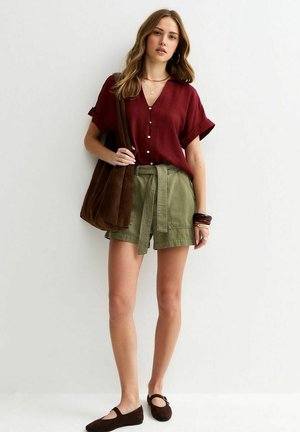 Kobieta ubrana w bordową bluzkę zapinaną na guziki, oliwkowozielone szorty z paskiem, brązowe baleriny, niosąca dużą brązową torbę na ramię na jednolitym tle.