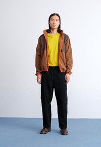 Chaqueta con cremallera marrón y naranja sobre una camiseta amarilla, combinada con pantalones negros y zapatos marrones. Tejido suave con un ajuste relajado.