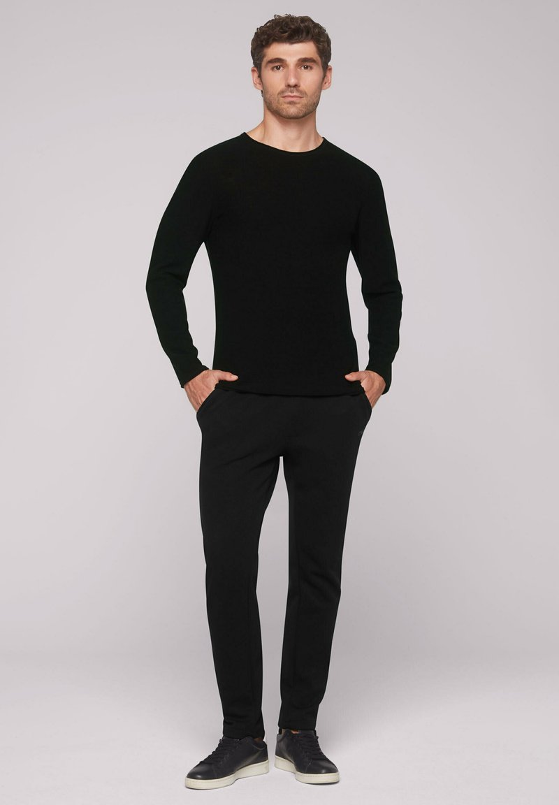 Schwarzes Langarmshirt, schlanke Passform, kombiniert mit schwarzer Hose. Das Model steht mit den Händen in den Taschen und trägt dunkle Sneaker. Glatte Textur.