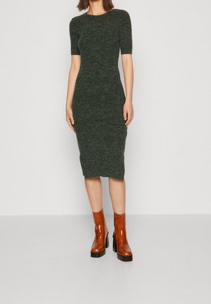 Kvinde iført en tætsiddende, ribstrikket mørkegrøn midi-kjole med korte ærmer og brune laklæder ankelstøvler med blokhæl.