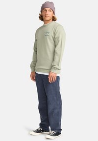 Lichtgroene sweatshirt met een golfgrafiek op de borst, gecombineerd met marineblauwe corduroy broek en zwarte sneakers met witte zolen.