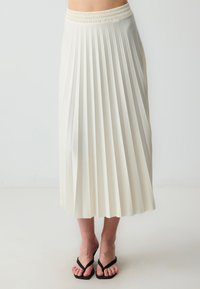 Plissé midi-rok in crème, gemaakt van lichte stof. Heeft een elastische tailleband en valt tot halverwege het scheenbeen. Gepaard met zwarte teenslippers.