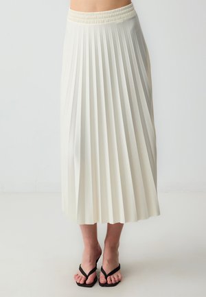 Faltenrock in Creme, aus leichtem Stoff hergestellt. Mit einem elastischen Bund und reicht bis zur Mitte der Wade. Kombiniert mit schwarzen Flip-Flops.