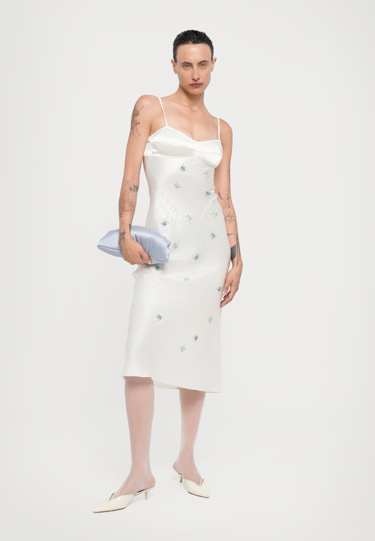 Robe nuisette en satin blanc avec des fines bretelles et une broderie florale bleue, assortie à une pochette bleu clair et des mules pointues crème.