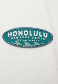 Patch ovale teal con testo in rilievo bianco "HONOLULU NEWPORT STORE CUSTOM BRANDS" e grafiche di onde. Bordo cucito in blu scuro.