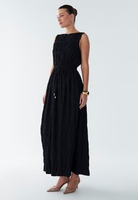 Schwarzes Maxikleid mit plissierter Textur, ärmellosem Design und Tunnelzug in der Taille. Kombiniert mit beigefarbenen, verzierten Absätzen und minimalistischem Schmuck.