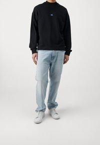 Hombre vestido con un sudadera negra de manga larga con un pequeño logo, jeans de mezclilla azul claros y zapatillas blancas, de pie frente a un fondo blanco liso.