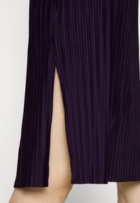 Jupe midi plissée en violet foncé, avec des plis verticaux, une fente latérale et une finition en tissu lisse et texturé.