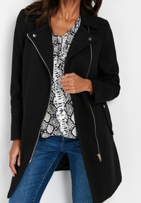 Černý kabát na zip s výstřihem s klopami a stříbrným kováním, nošený přes vzorované tričko, doplněný modrými džínami. Měkká, texturovaná látka.