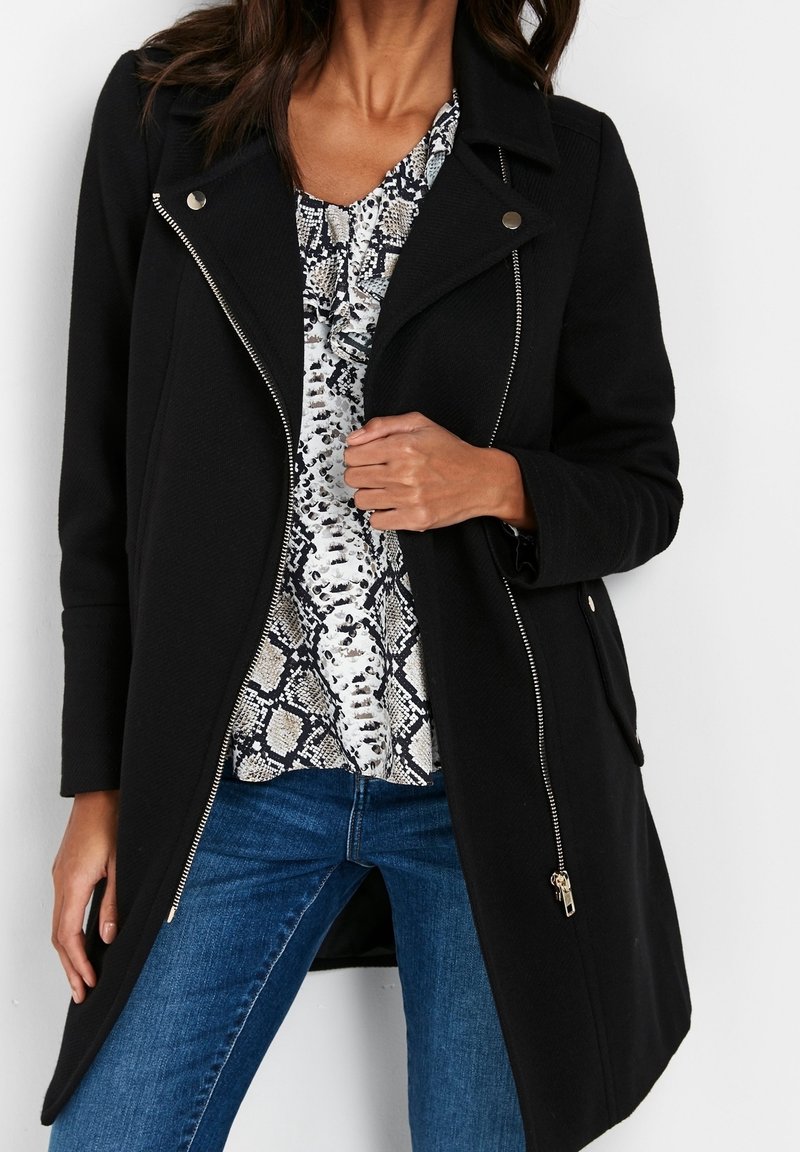 Černý kabát na zip s výstřihem s klopami a stříbrným kováním, nošený přes vzorované tričko, doplněný modrými džínami. Měkká, texturovaná látka.