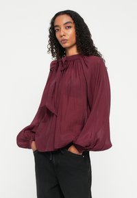 Femme portant un chemisier prune à manches longues avec un nœud au col et un pantalon noir, les mains dans les poches, sur un fond blanc uni.