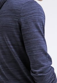 Pull en tricot bleu marine avec un motif texturé à rayures. Il comporte des manches longues et un col rond, avec des coutures visibles à la couture des épaules.