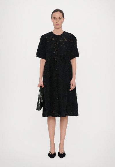 Black dress with textured floral pattern, short sleeves, and a relaxed fit, paired with a black croc-embossed bag and pointed black shoes.