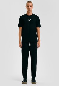 Zwart katoenen t-shirt met een klein grijs logo op de borst, gecombineerd met zwarte broek met een koord, gedragen met zwarte sneakers.