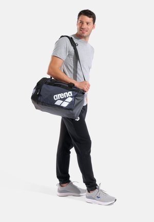 Uomo con maglietta grigia e pantaloni neri che porta una grande borsa da palestra nera Arena sulla spalla, in piedi e guarda di lato.