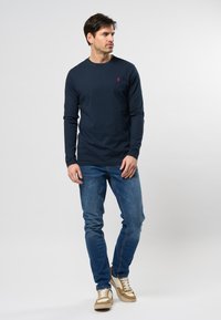 Camicetta a maniche lunghe blu navy con un piccolo logo rosso, abbinata a jeans azzurri e sneakers beige a più trame. Design semplice, forma aderente.