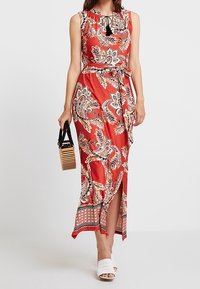 Abito maxi a fiori rossi con scollatura a fiocco, spacco laterale e vita rifinita da una cintura. Abbinato a sandali bianchi con tacco a blocco e una borsa a righe.