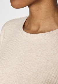 Lys beige struktureret stof med et ribbet mønster og en bred halsudskæring. Fokuser på skulder- og halsområdet. Glat, blød fremtoning.